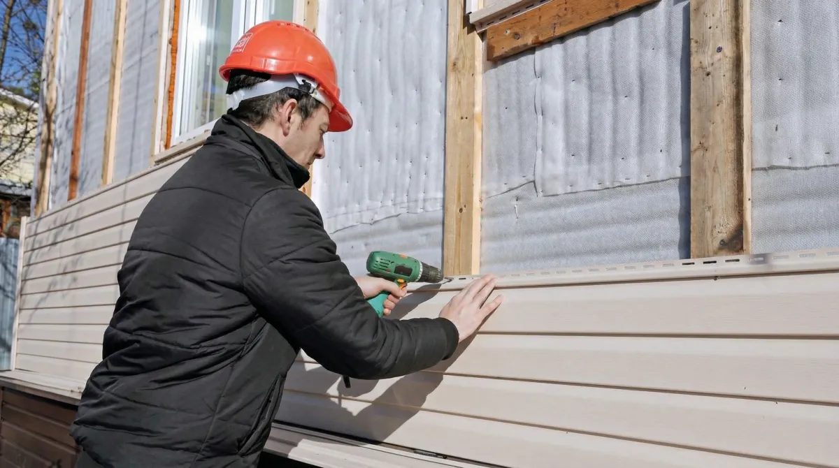 Siding replacement professional using a power drill to install new tan vinyl siding on a home exterior in Des Moines, IA